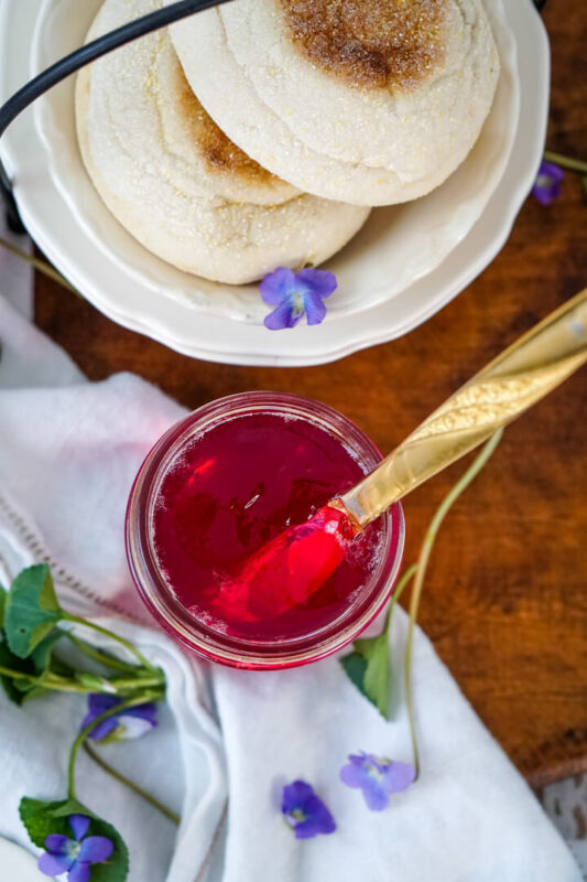 A jar of wild violet jelly with several flowers surrounding the jar and a plate to one side. 