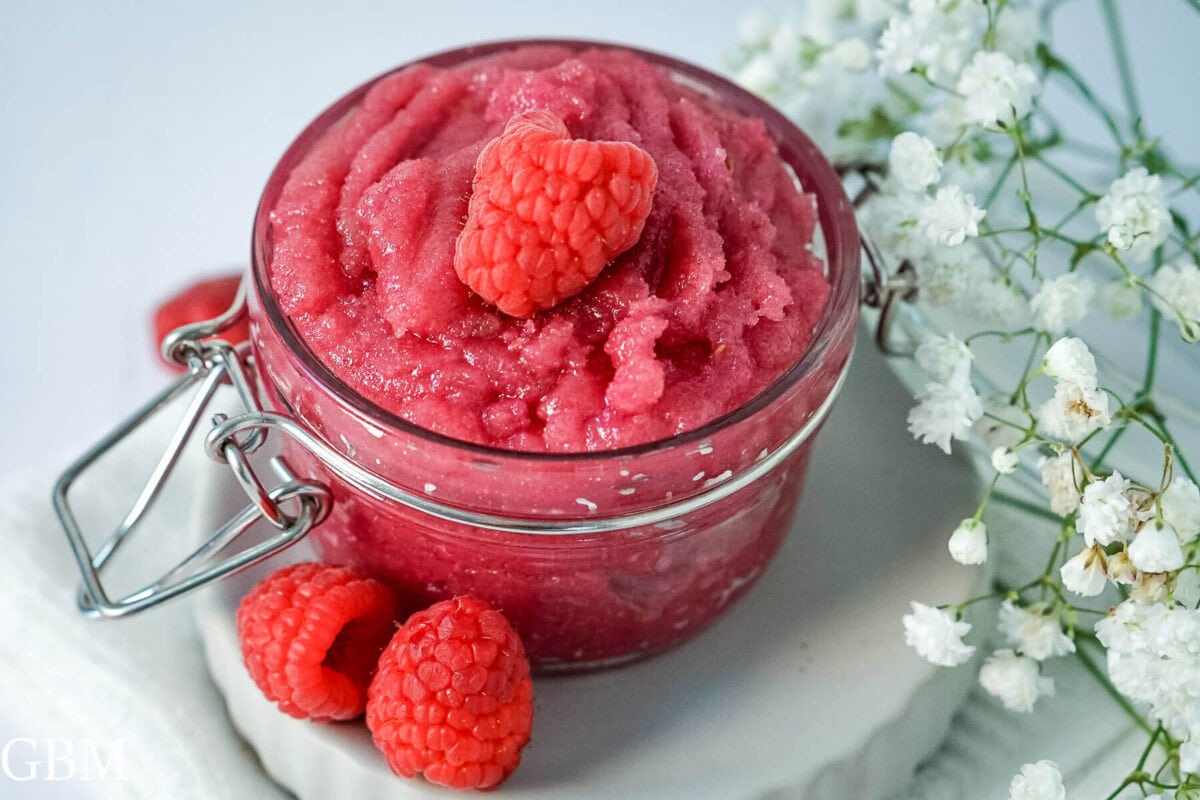 A jar of homemade raspberry sugar scrub with fresh raspberries to one side. 